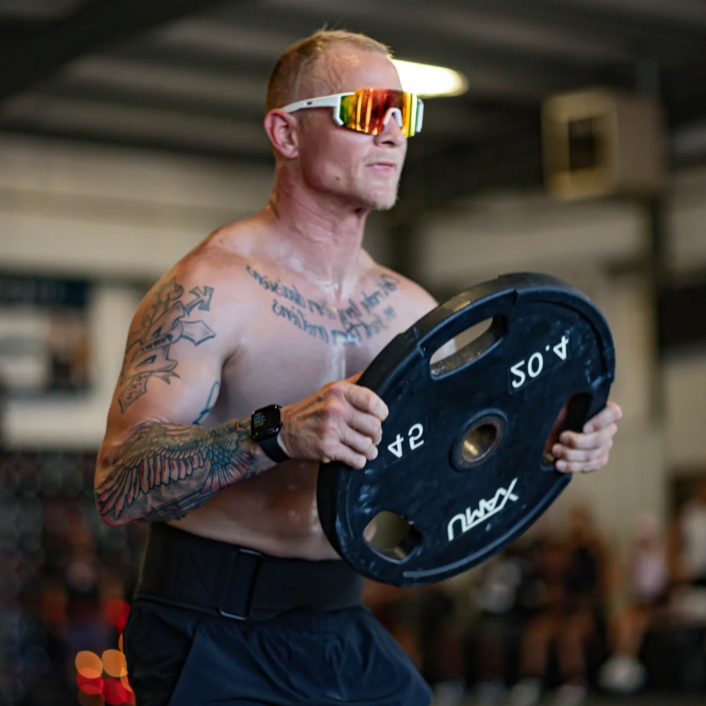 Tattooed athlete wearing sunglasses carrying a 45 lb weight plate during a True Grit Fitness class in Edmond, Oklahoma